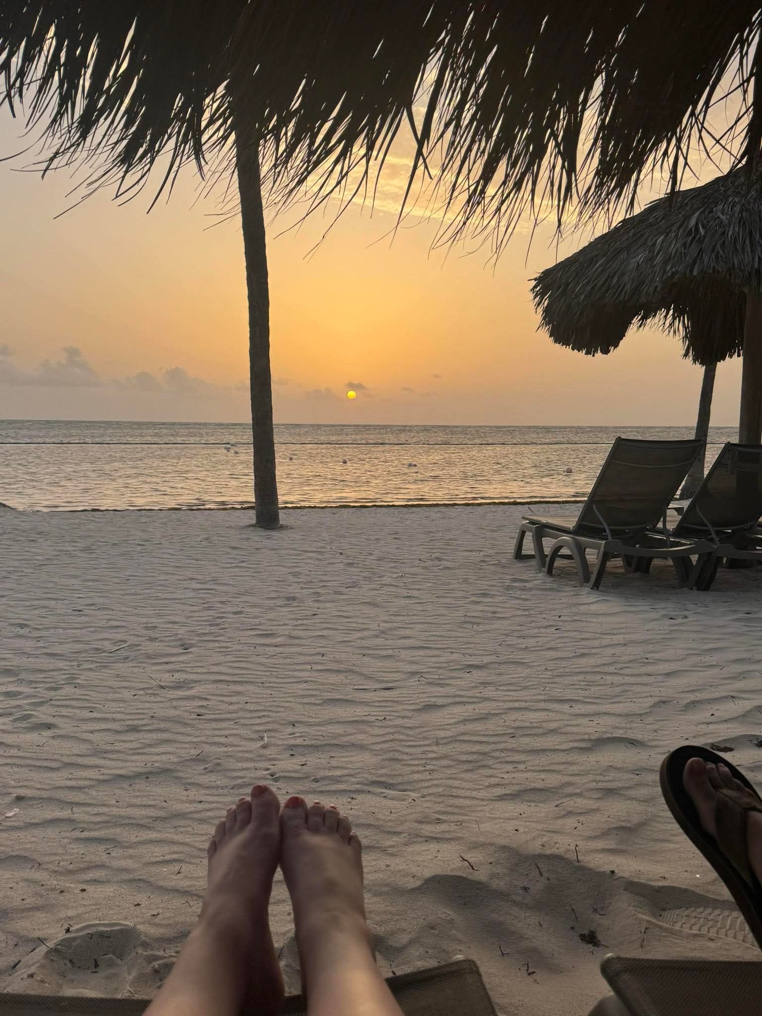 Beach sunset view with feet in foreground, lounge chairs, and thatched umbrellas.
