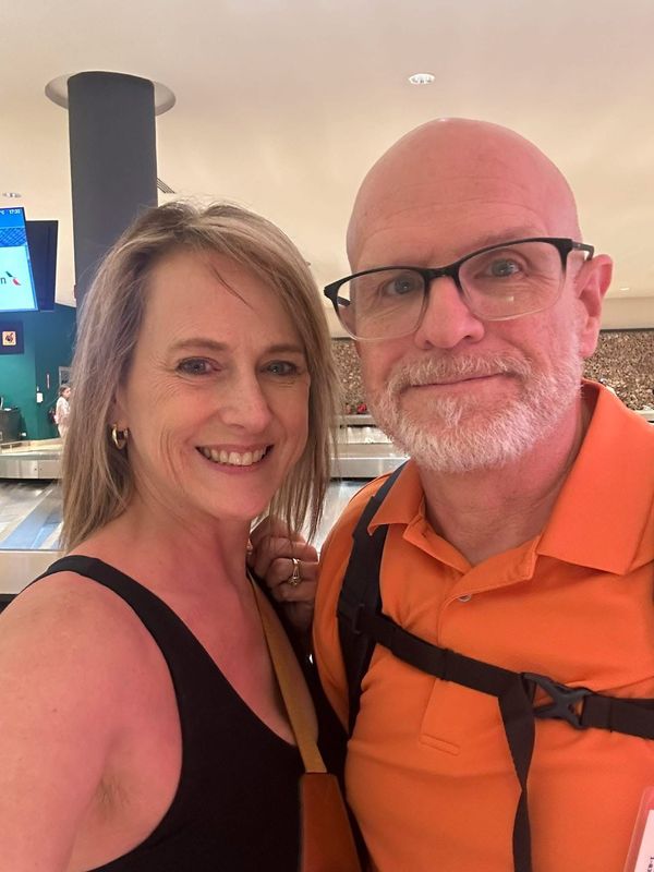 A smiling woman in a black tank top and a smiling man with glasses in an orange polo shirt pose together indoors.