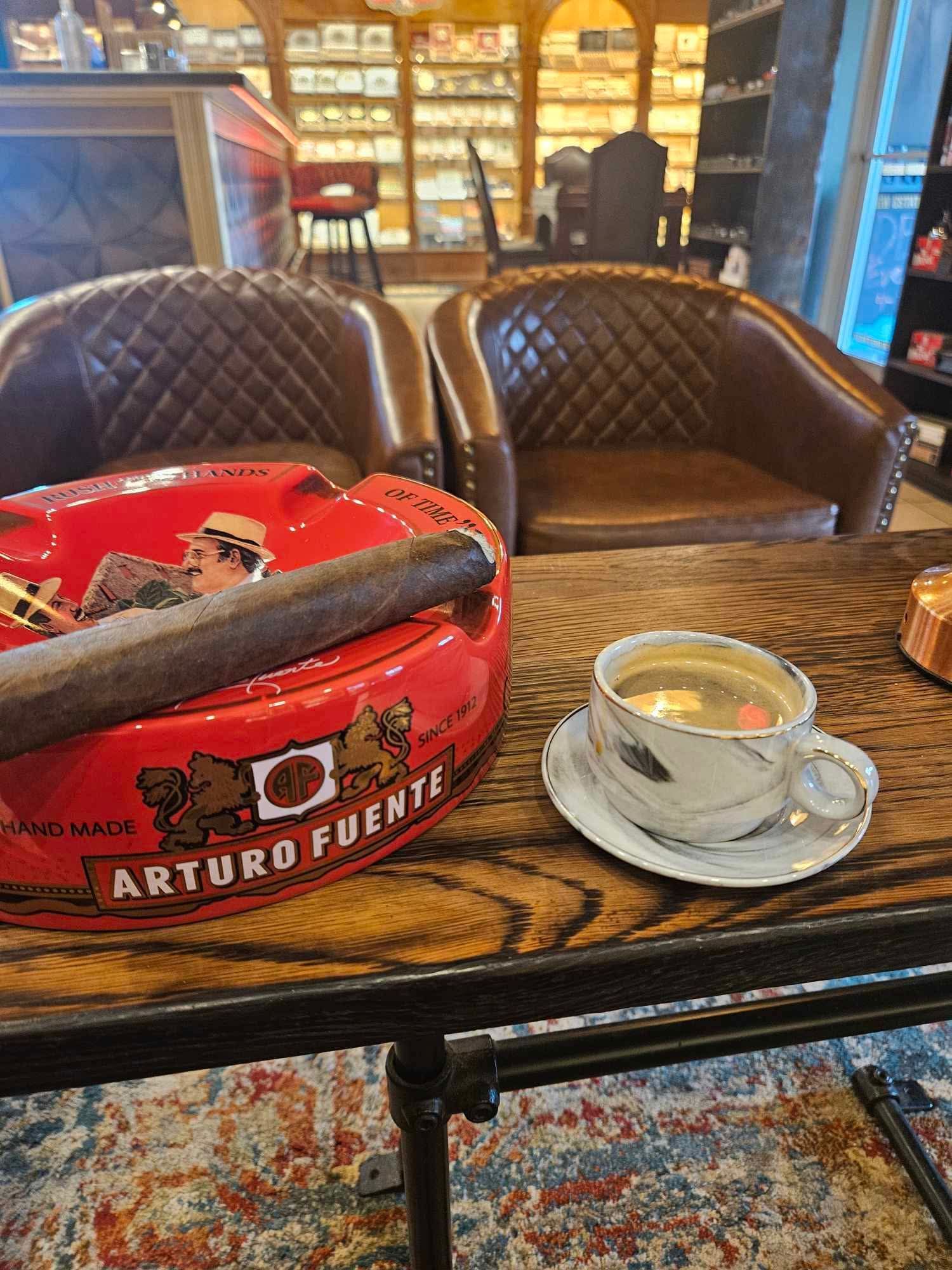 Cigar on Arturo Fuente ashtray next to coffee cup on a wood table, two leather chairs in the background.