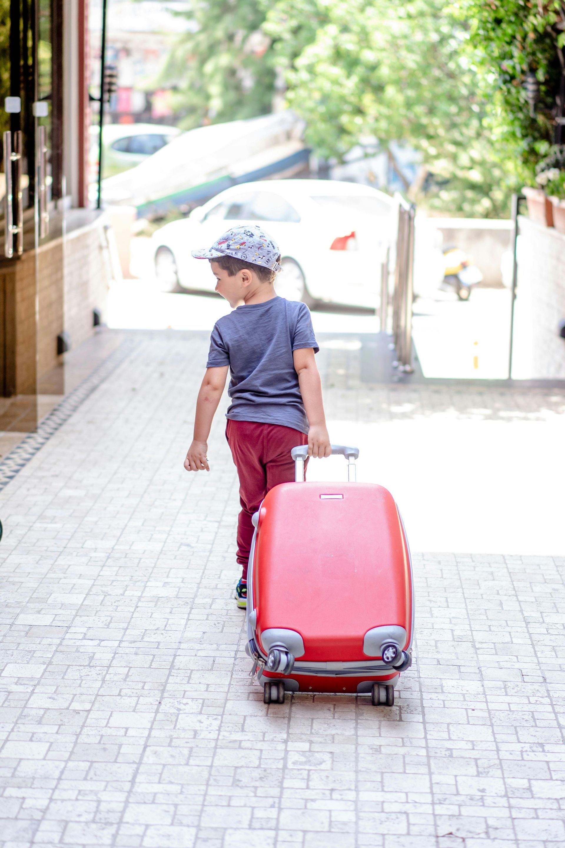 Child pulling a red suitcase along a sunlit sidewalk