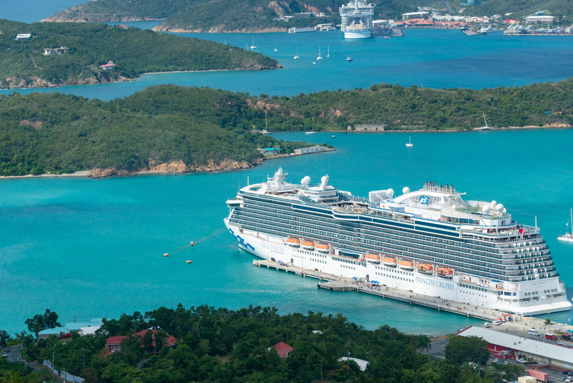 A large white cruise ship docked at a pier in a turquoise bay, surrounded by green tropical islands.