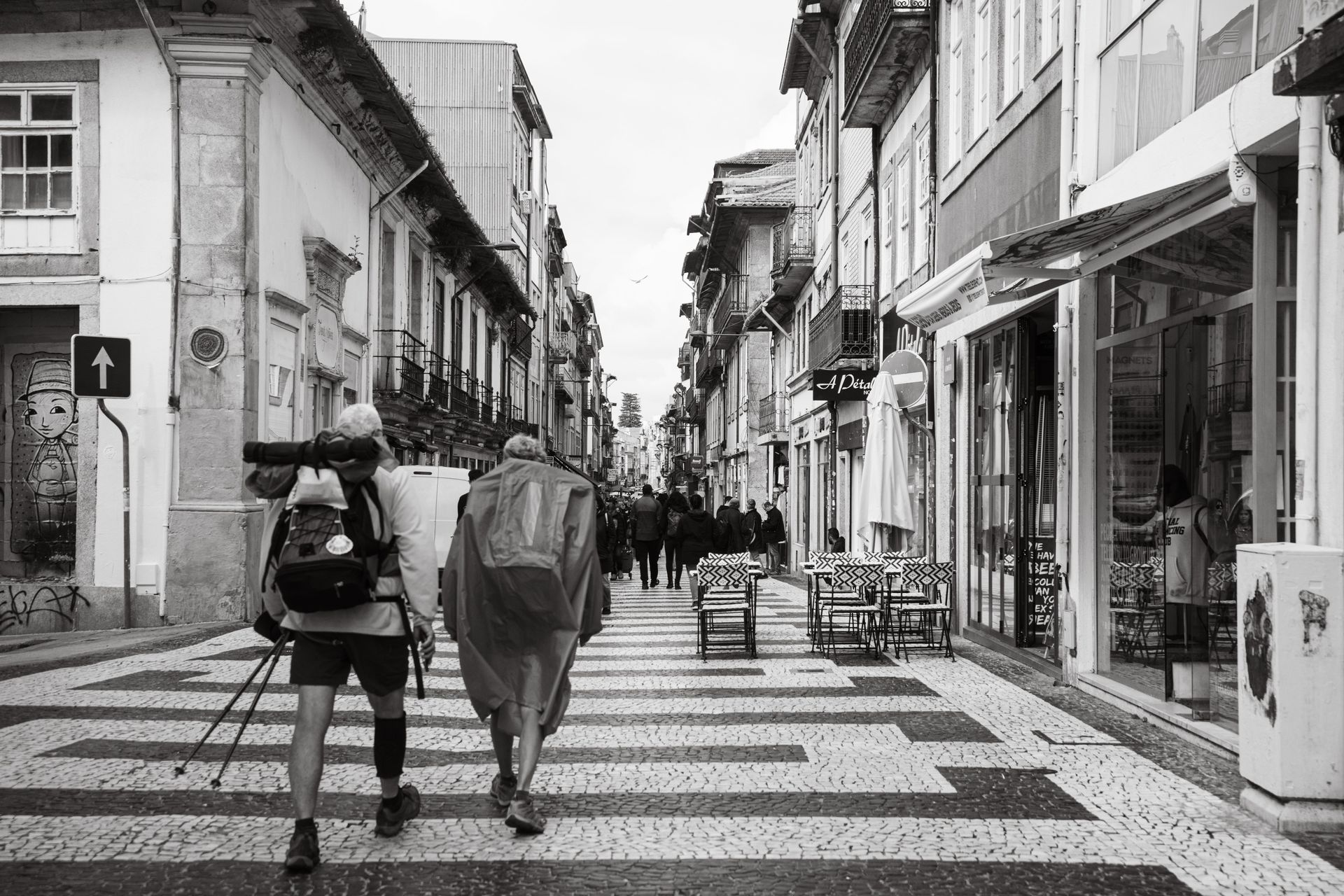 Two people with backpacks crossing a zebra crossing on a narrow city street with shops and buildings