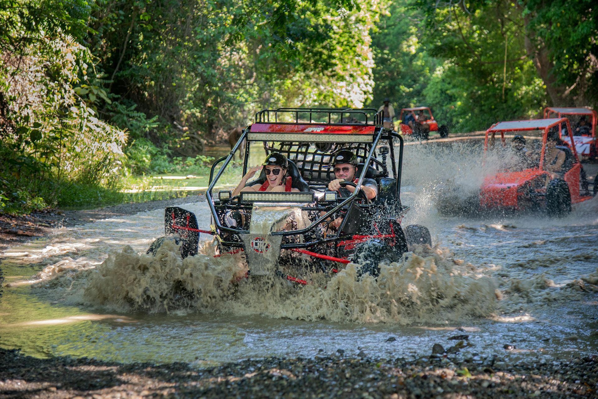 Two people in an off-road buggy splashing through a muddy forest trail, with another buggy following behind.