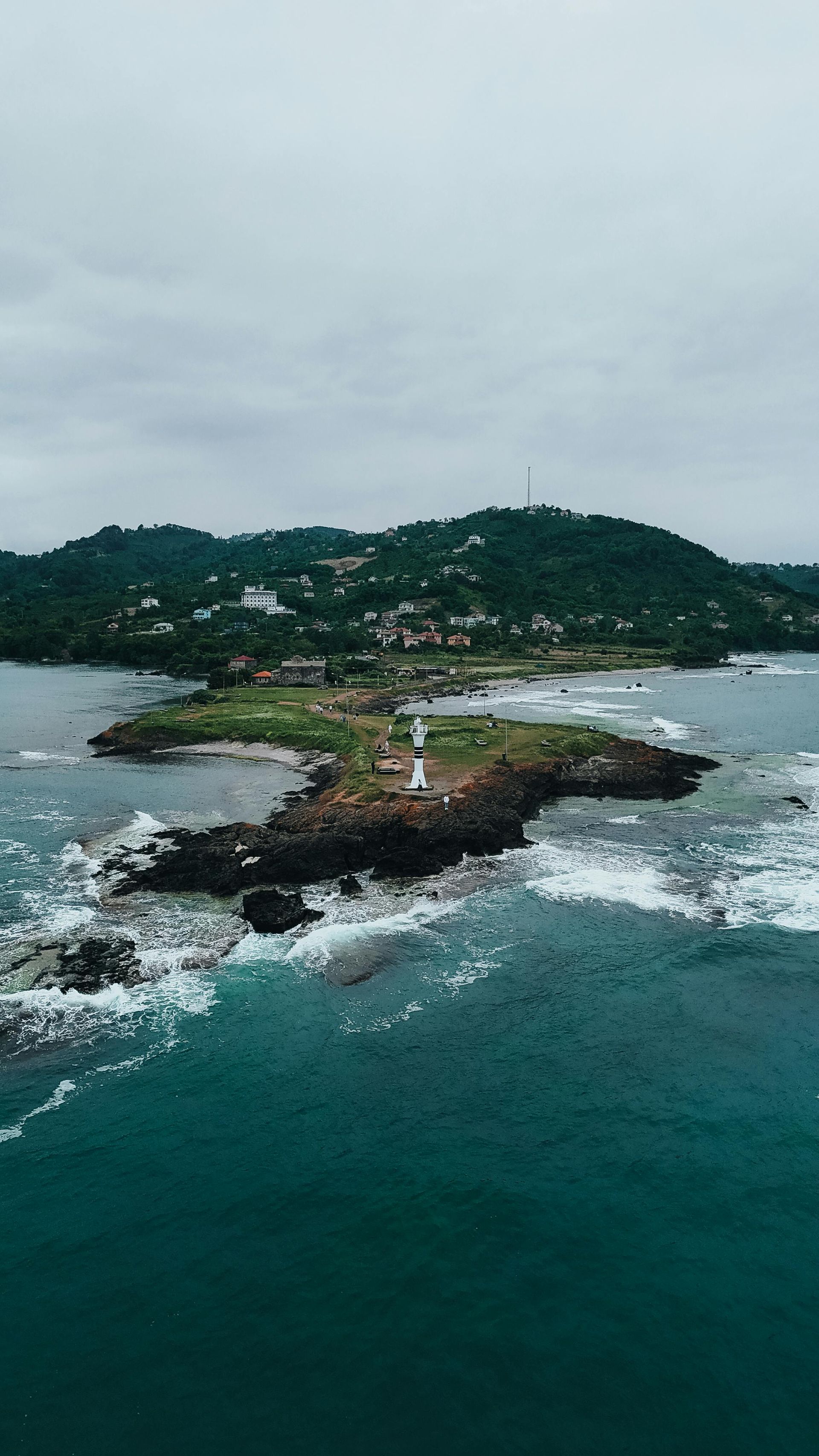 Rocky island with a small lighthouse, surrounded by teal water and white surf under a cloudy sky
