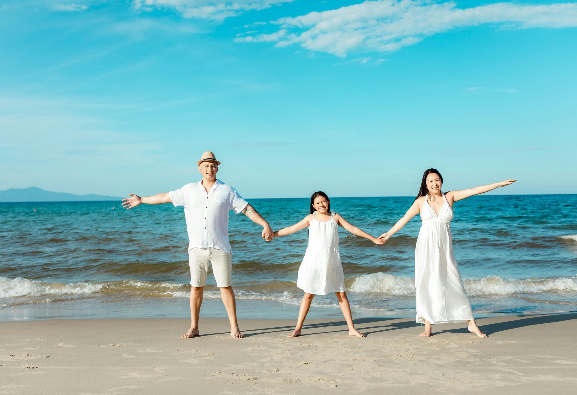 Three people in white clothing hold hands with arms outstretched on a sunny beach, standing before the blue ocean.