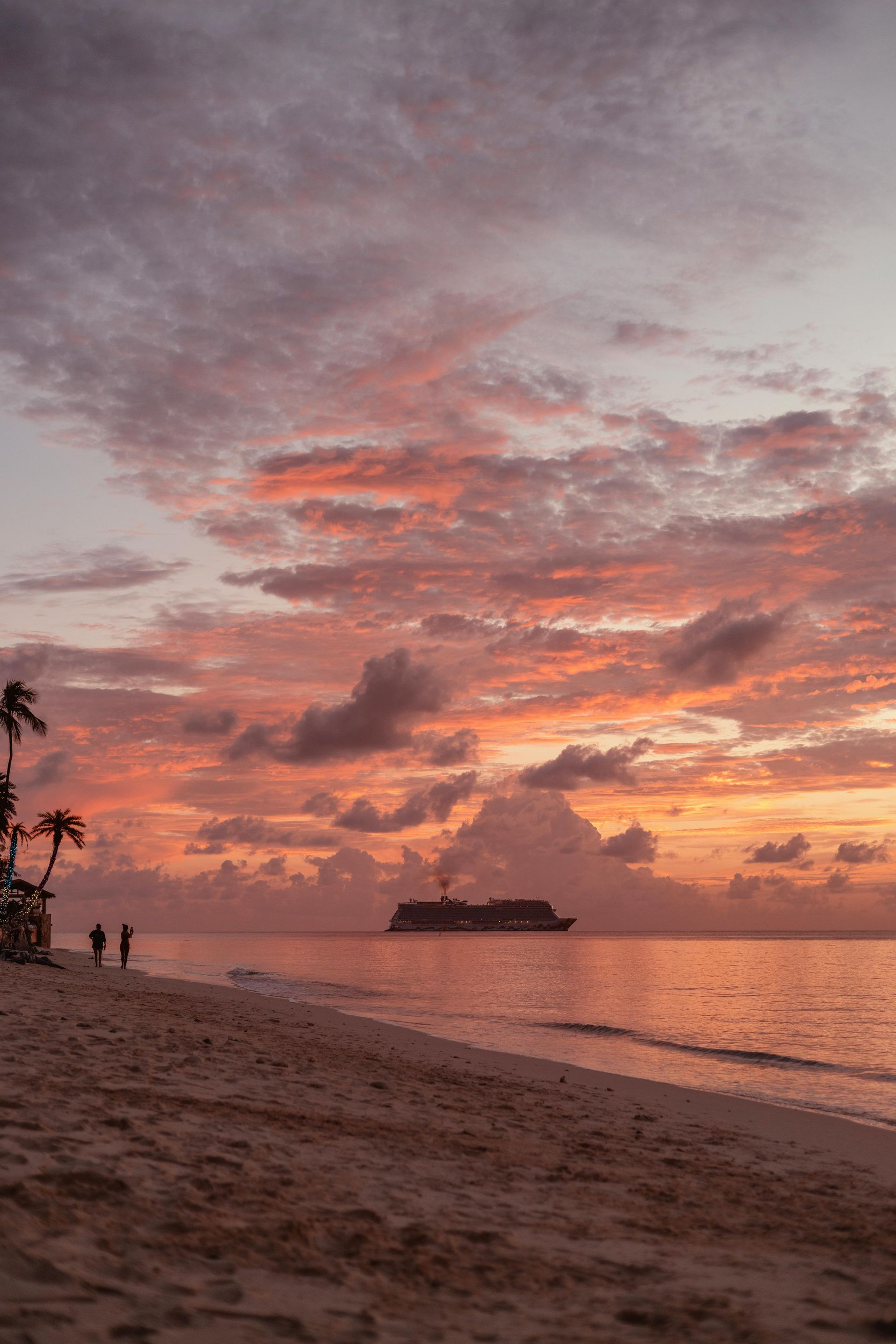 Beach at sunset with pastel sky, cruise ship on water, and silhouetted figures.