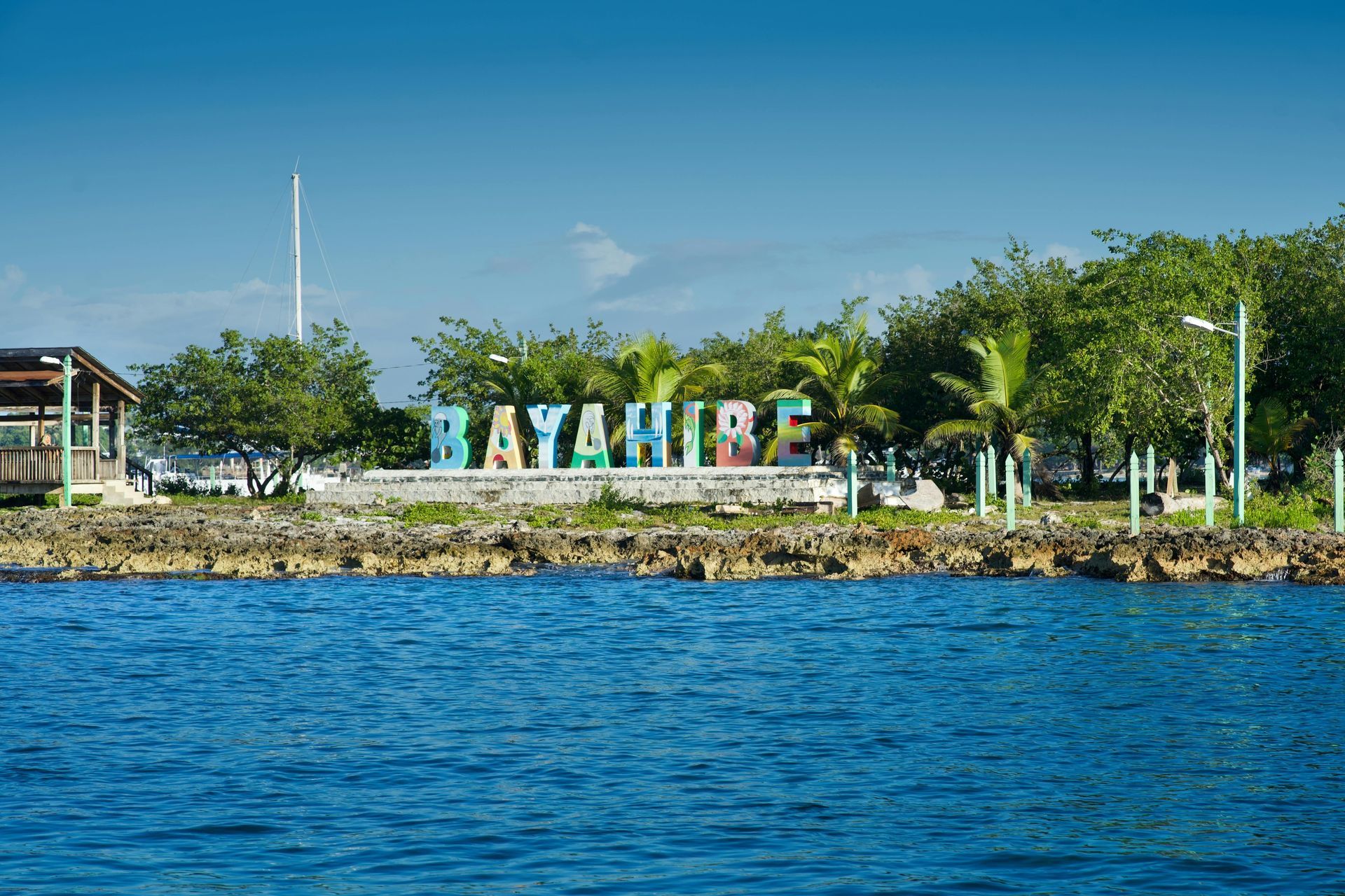 Colorful BAYAHIBE sign on a rocky shoreline beside blue water under a clear sky