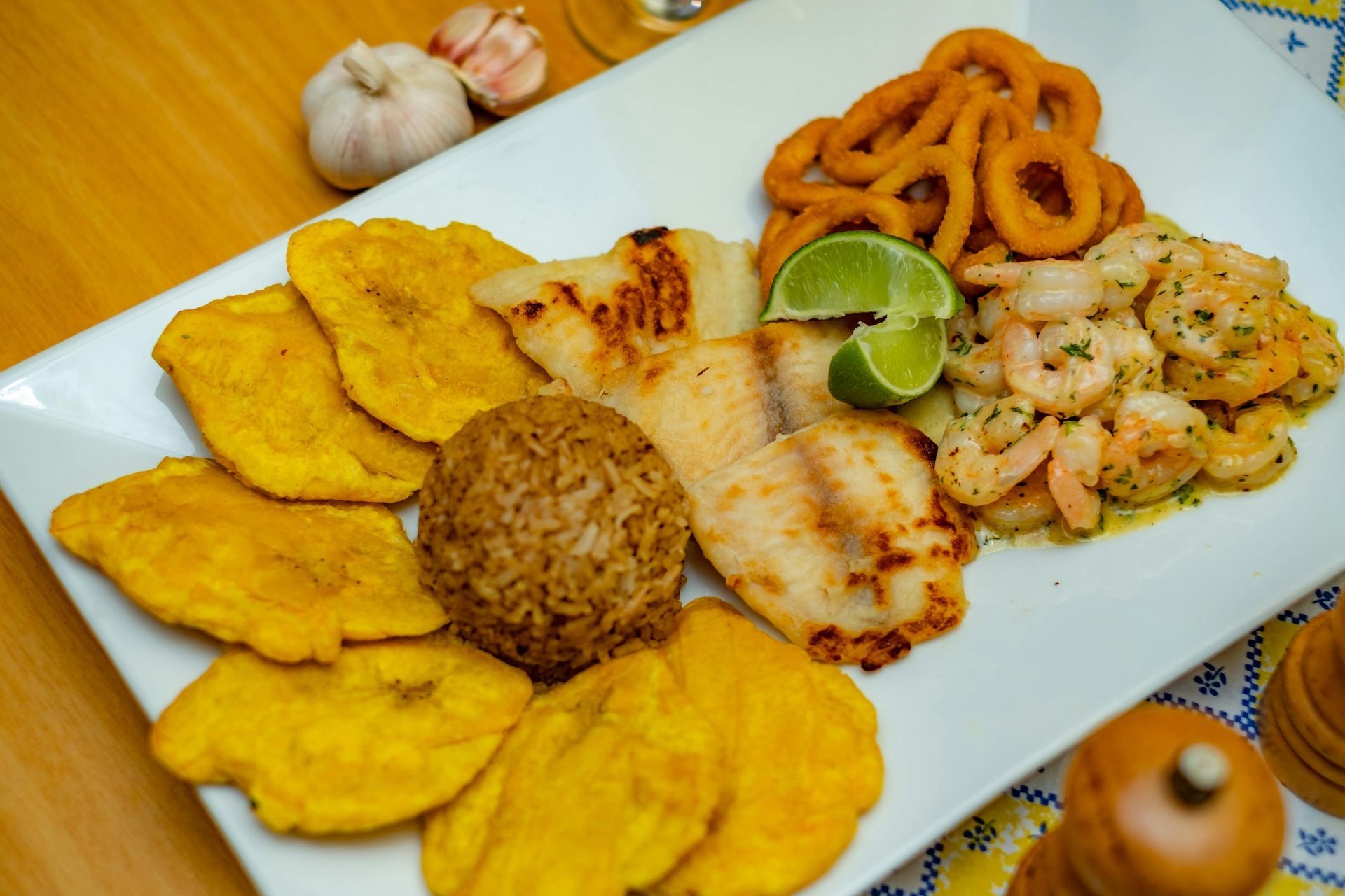 Plated seafood meal with fried plantains, rice, grilled fish, shrimp, lime, and fried calamari on a white dish