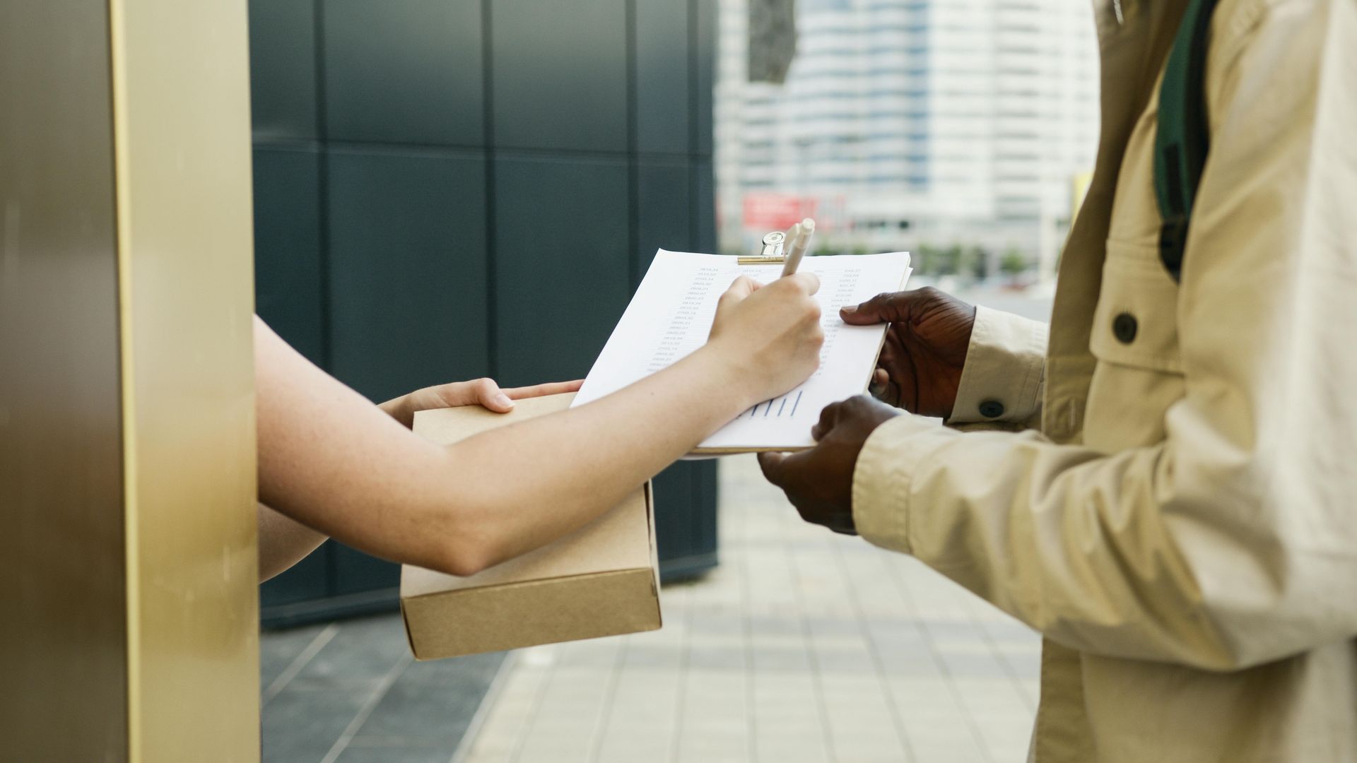 Hands exchanging a clipboard at a storefront counter with a cardboard package nearby.