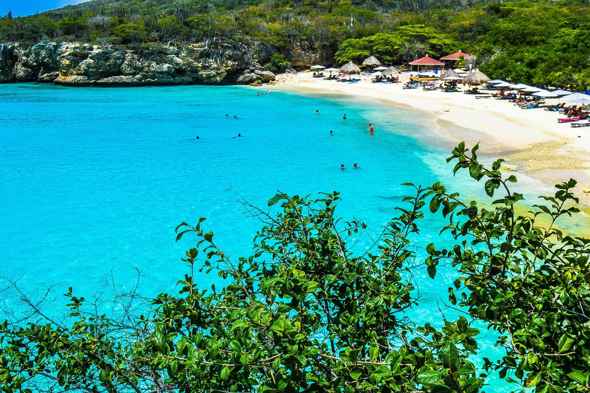 A scenic, turquoise bay with a sandy beach, surrounded by lush green cliffs and foliage, under a bright sunny sky.