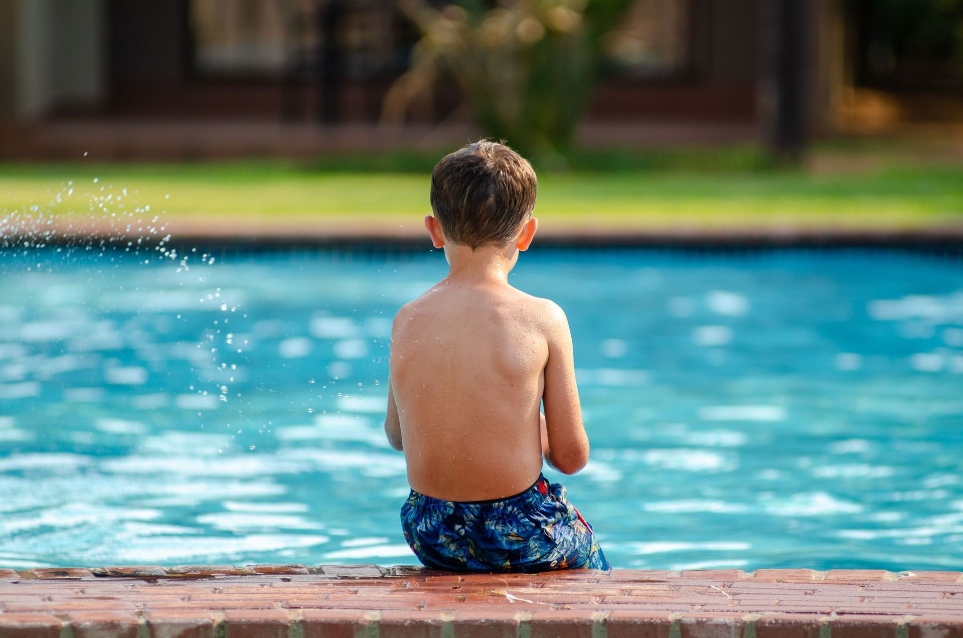 Child sitting on a pool edge with back to camera, wearing blue swim trunks, facing the water