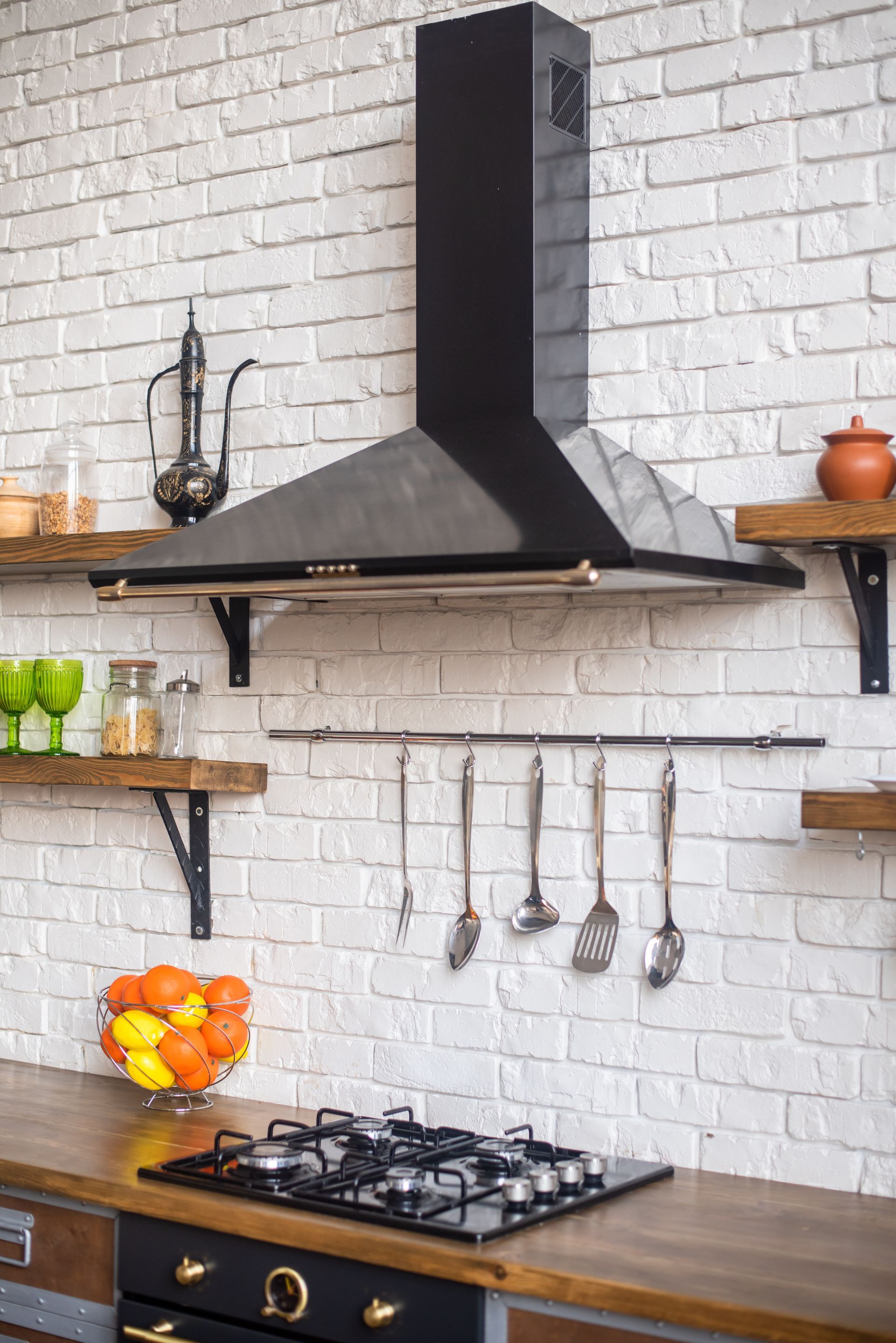 A kitchen with a stove top oven , a hood , shelves and a brick wall.