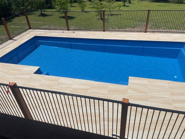 Blue tiled swimming pool surrounded by a beige tiled deck and black metal fence.