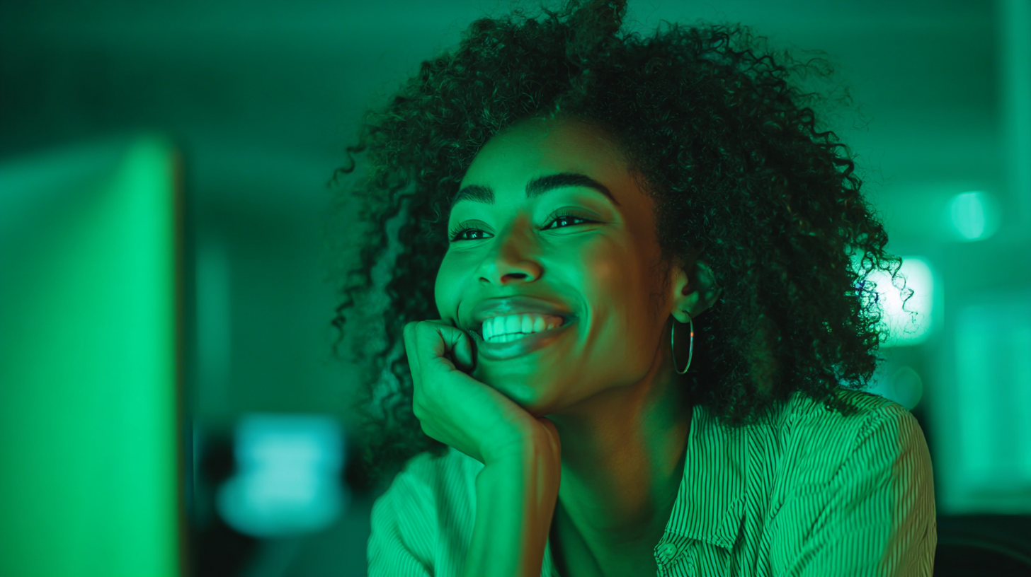Uma mulher de cabelo cacheado sorri enquanto olha para a tela de um computador em uma sala iluminada