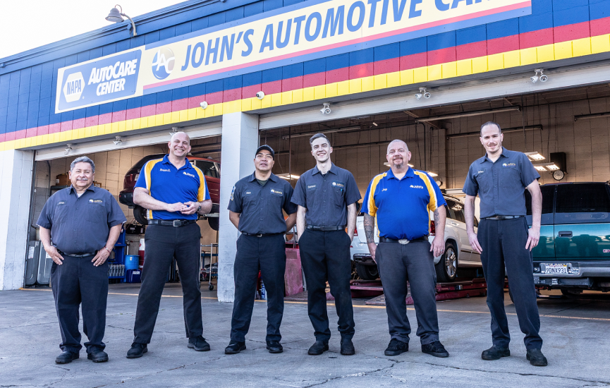Six automotive technicians in blue/gray shirts stand in front of John's Automotive Car Repair shop.