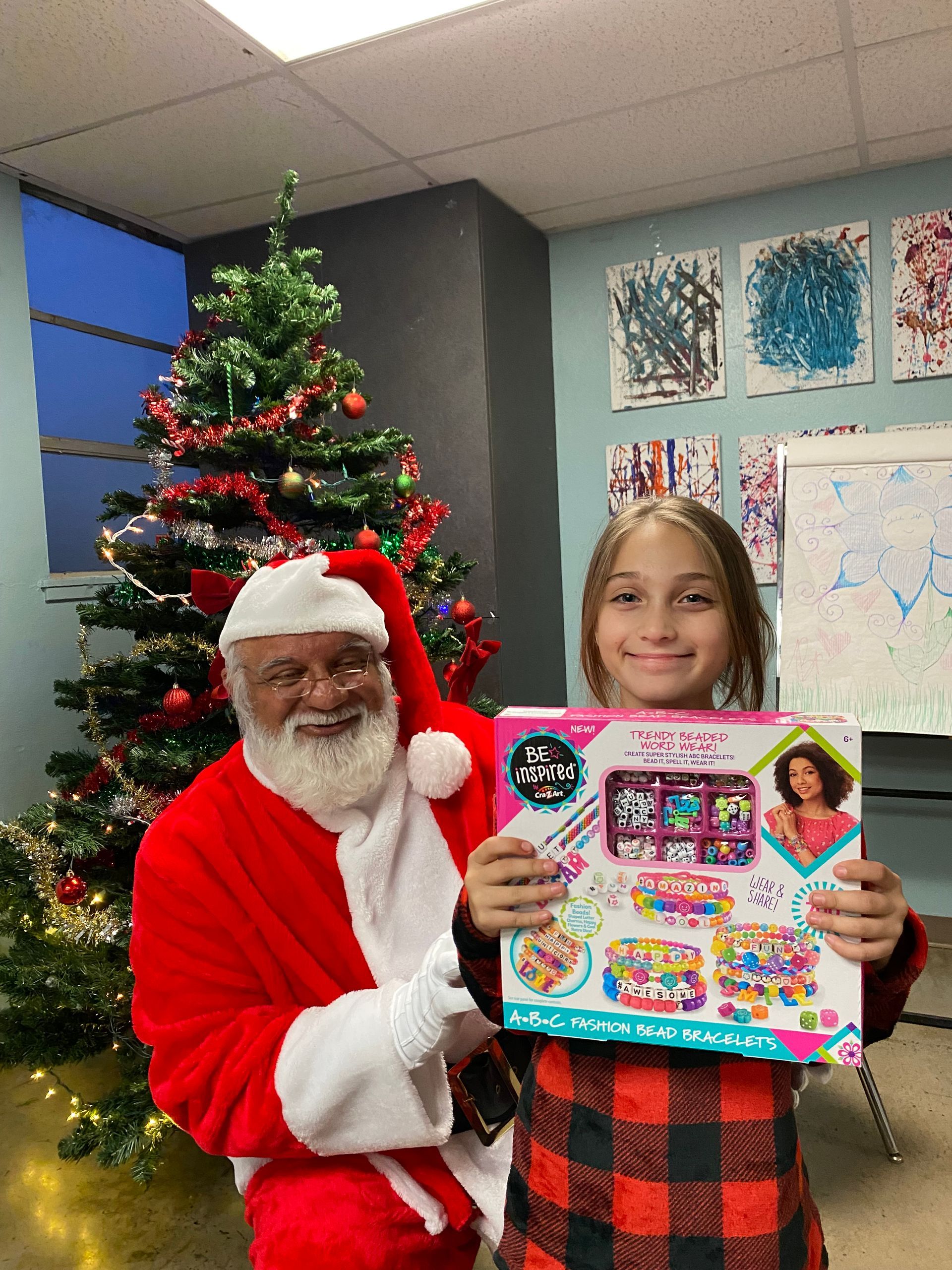Santa Claus with a young person, holding a gift, in front of a Christmas tree.