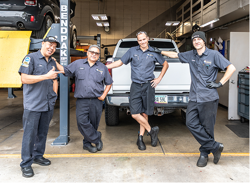 Four auto mechanics pose in a garage, smiling. A truck is behind them on the lift.