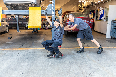 Two mechanics playing in a garage: one on a creeper with arms up, the other pushing him with a smile.