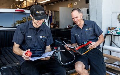 Two people in work shirts reviewing papers in a truck bed, one holding a book, both smiling.