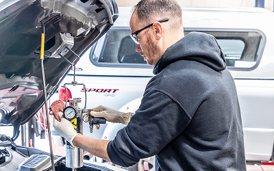 Mechanic working on a car engine in a shop, using tools. The hood is open, and a white truck is in the background.