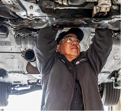Mechanic working under a car, wearing a cap and jacket, in a garage.