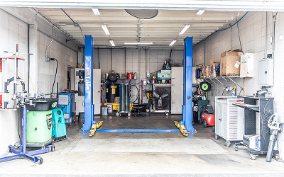 Interior of a garage with a car lift, tools, and equipment. Blue lift, gray walls, overhead lights.