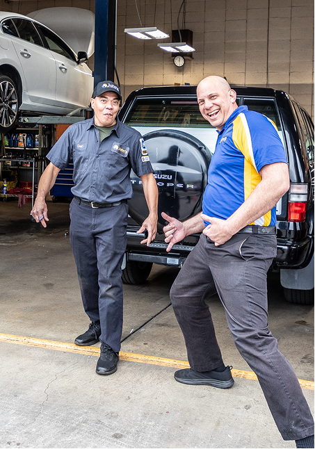 Two men in a garage, one points, the other smiles with a hand gesture near a black SUV; car on a lift in the background.