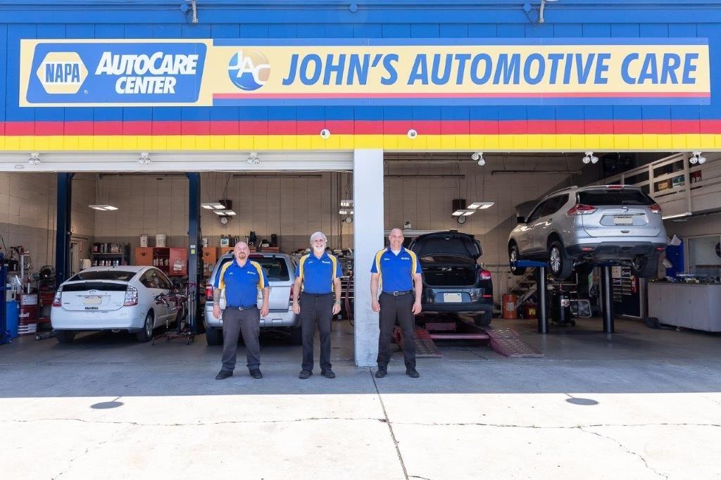 Three auto shop employees in blue shirts stand in front of John's Automotive Care. Cars are in bays.