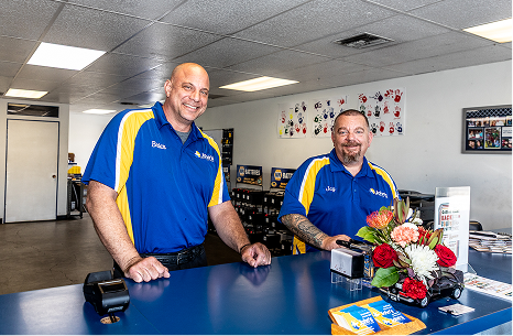 Two men behind a counter in blue and yellow shirts, smiling at the camera in a business setting.