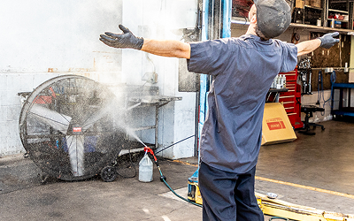 Man in work clothes sprays water into fan, arms outstretched, in a garage.