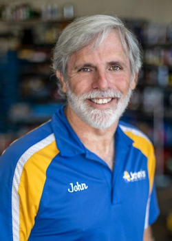 Man with gray beard smiles, wearing a blue and yellow shirt, in a garage.