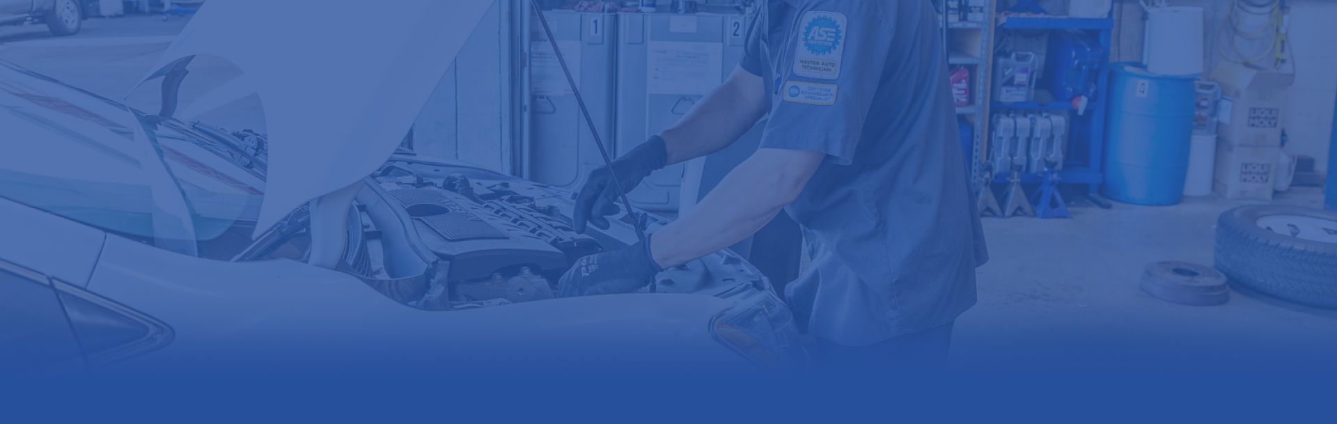 Mechanic with open mouth holding tools in a garage setting, wearing a work shirt.