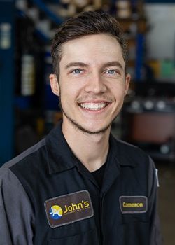Man in mechanic's uniform smiles at camera, posing at John's auto shop.