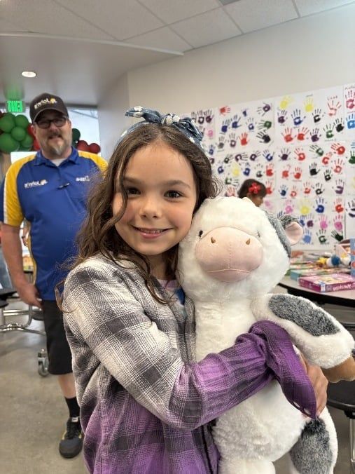 Girl smiles, holding a stuffed cow. Man in background, handprints on wall.