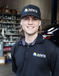 Man in John's Automotive uniform smiles, wearing a cap. He's inside a shop with shelves of parts in the background.