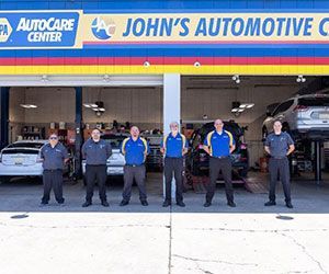 Group of six auto mechanics in uniform standing in front of a repair shop, John's Automotive.