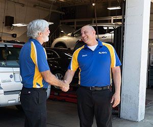 Two auto shop employees shaking hands. One has gray hair. They are in a garage with cars.