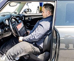 Mechanic in a car, using a diagnostic tool. Gray car, dark-haired man in blue shirt, focused expression.