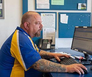 Man with tattoos typing at a computer, wearing a blue and yellow shirt. Office setting with bulletin board.