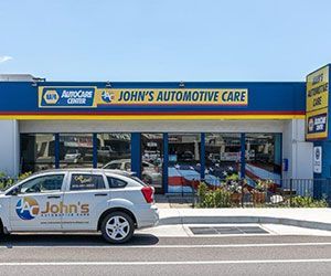 John's Automotive Care shopfront with blue and yellow signage, a car in the foreground, and cars visible inside.