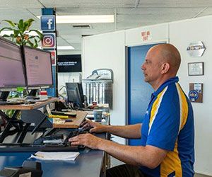 Man working at a desk with two monitors; blue and yellow shirt, typing. Social media logos on the wall.