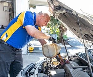 Mechanic pouring oil into a car engine, wearing a blue and yellow shirt, outdoors.