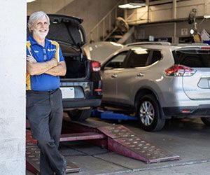 Mechanic in blue shirt, arms crossed, leans in garage. Silver SUV on lift behind.