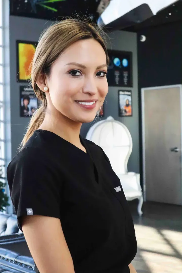 Woman in black scrubs smiles, posing near a decorative chair indoors, with artwork on the wall.