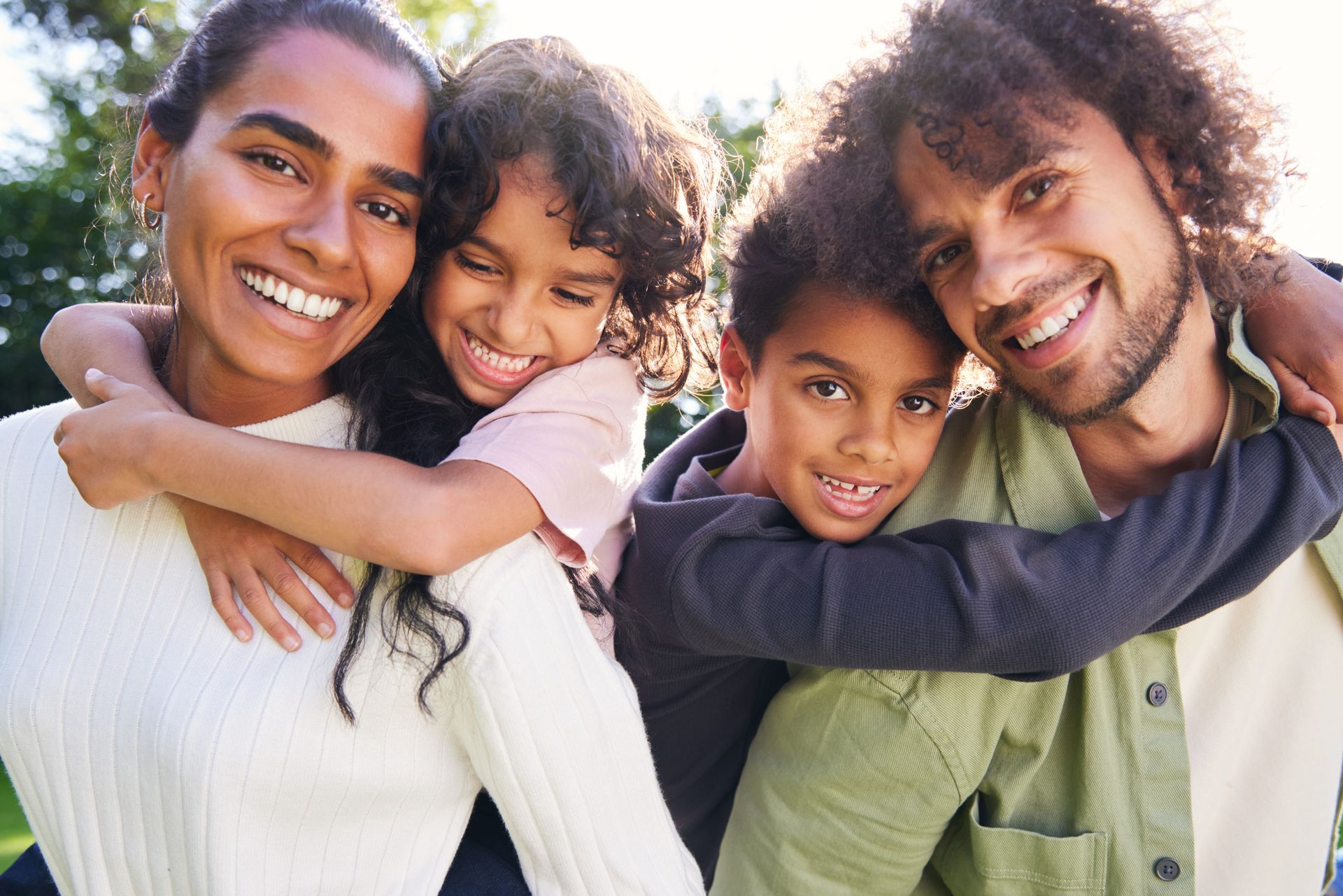 Family of four smiling, embracing outdoors. Parents with two children. Sunlight, green background.