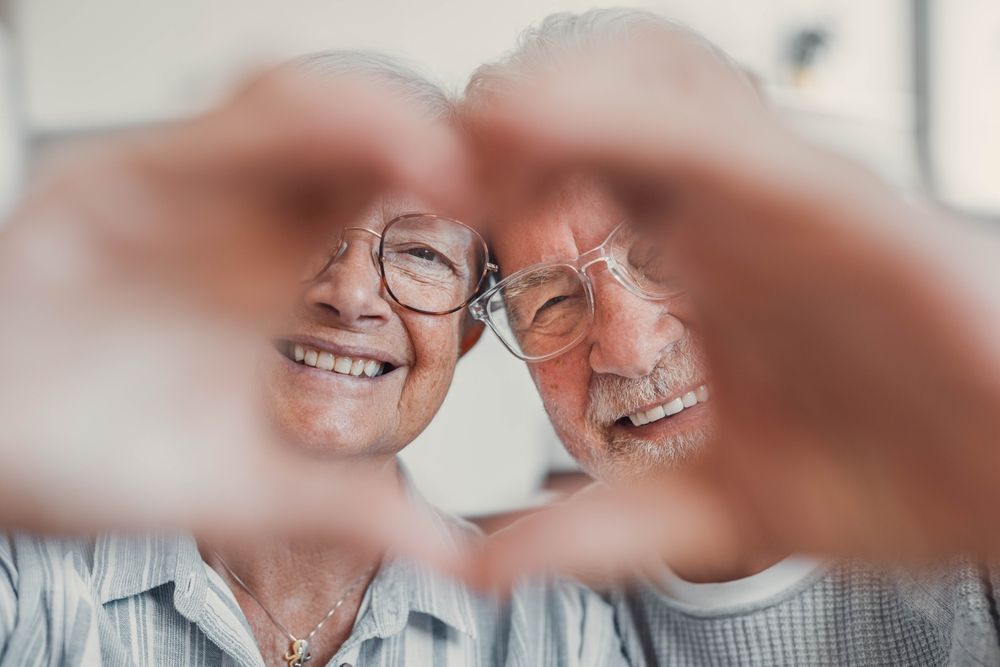 Smiling couple framing their faces with hands, wearing glasses, in a bright indoor setting
