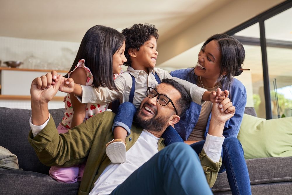 Family playing on a couch, smiling and holding hands in a bright living room.