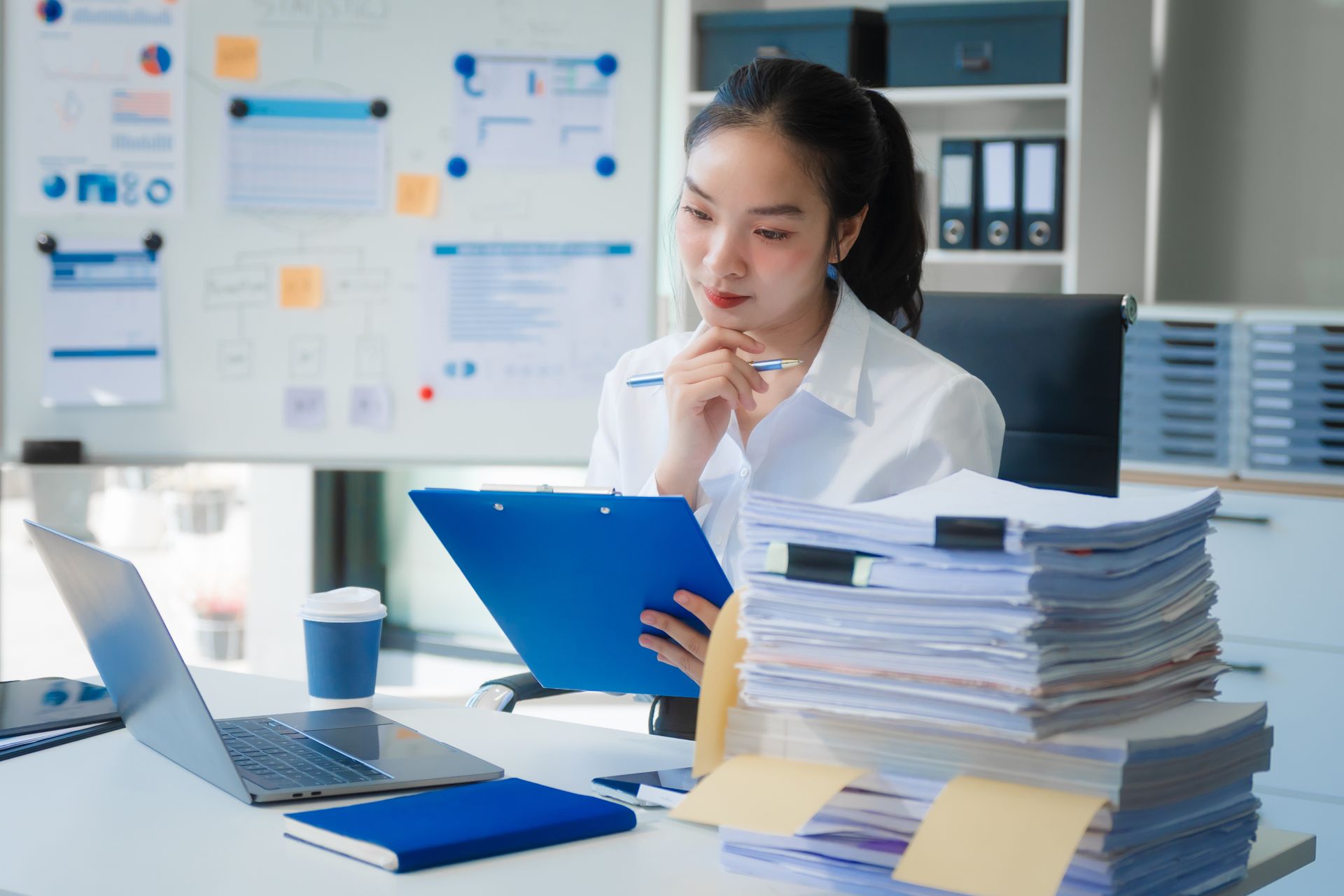 A woman is sitting at a desk with a laptop and a stack of papers.