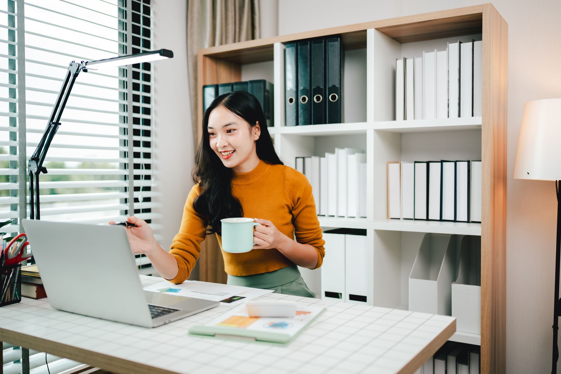 A woman is sitting at a desk using a laptop computer while holding a cup of coffee.