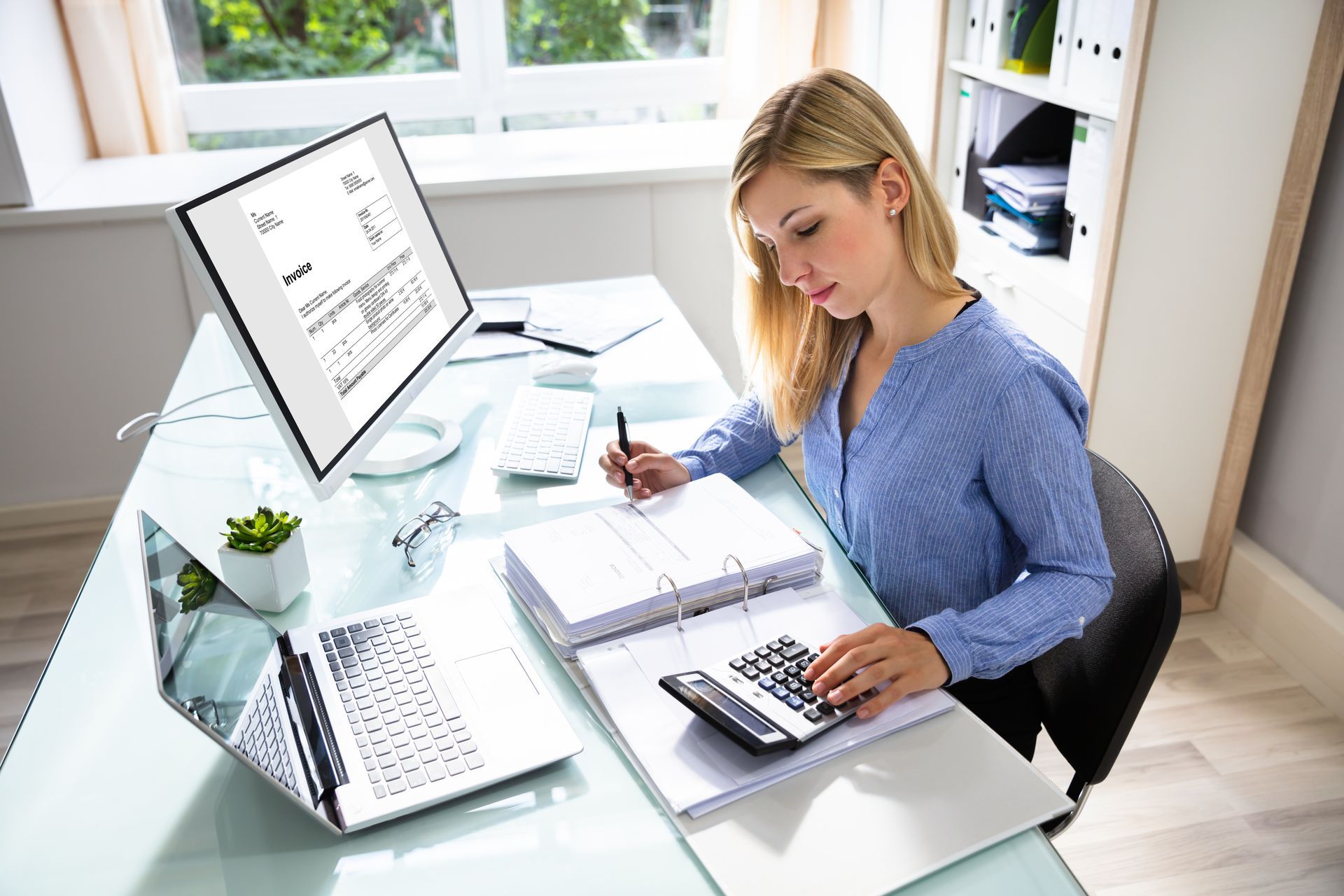 A woman is sitting at a desk using a computer and a calculator.